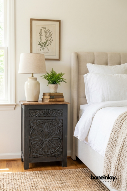 Handcrafted black carved solid wood bedside cabinet with intricate floral medallion door, natural wood top, single door storage, and traditional hand-carved detailing, styled beside an upholstered bed in a modern farmhouse bedroom.