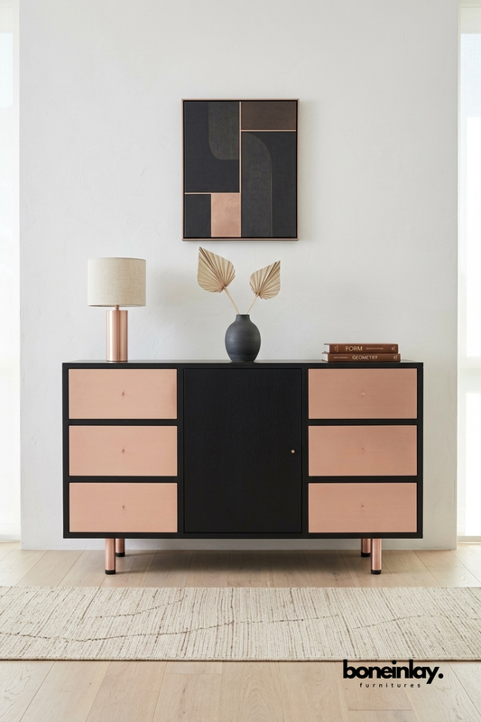 Black and copper two-tone sideboard featuring six blush copper drawer fronts and a central black cabinet door. Slim copper legs add a refined Art Deco-inspired silhouette to the design. Styled with a copper table lamp, dried palm fronds, and stacked books beneath framed geometric wall art. A bold statement sideboard for modern and glam interior styling.