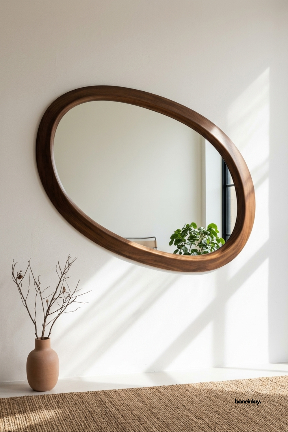 Hand-carved organic oval wooden mirror styled in a sunlit minimalist entryway alongside a terra-cotta vase and a woven jute rug. Featuring a smooth contoured walnut frame, this chic decorative wall mirror brings natural warmth, sculptural art, and mid-century modern sophistication to modern homes.