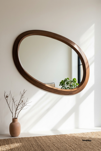 Hand-carved organic oval wooden mirror styled in a sunlit minimalist entryway alongside a terra-cotta vase and a woven jute rug. Featuring a smooth contoured walnut frame, this chic decorative wall mirror brings natural warmth, sculptural art, and mid-century modern sophistication to modern homes.