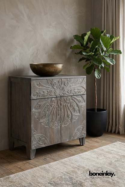 Grey-washed wood cabinet with a large carved floral medallion design.
One drawer sits above a two-door cabinet base for storage.
Whitewashed detailing highlights the petal and leaf carving.
Styled beside a fiddle leaf fig plant and brass bowl.