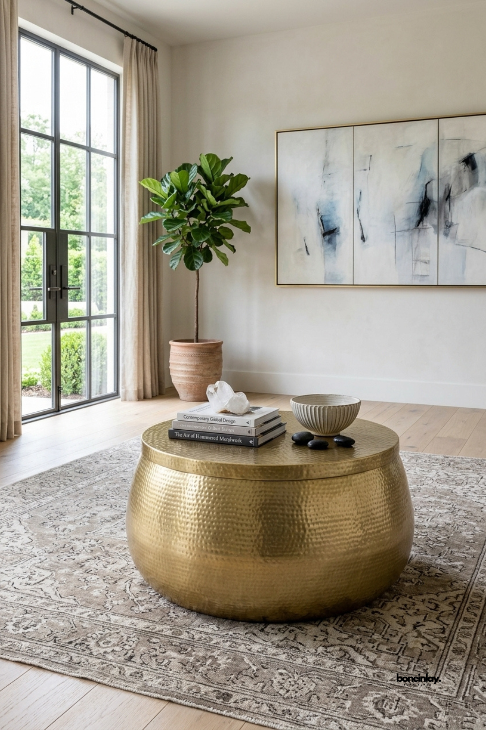Handmade round brass drum coffee table featuring a textured hammered metal finish and a smooth flat tabletop. Styled in a bright modern living room on a vintage Persian rug with an olive tree and abstract wall art, this artisan metallic cocktail table brings warm luxury, tactile elegance, and bohemian sophistication to contemporary home interiors.