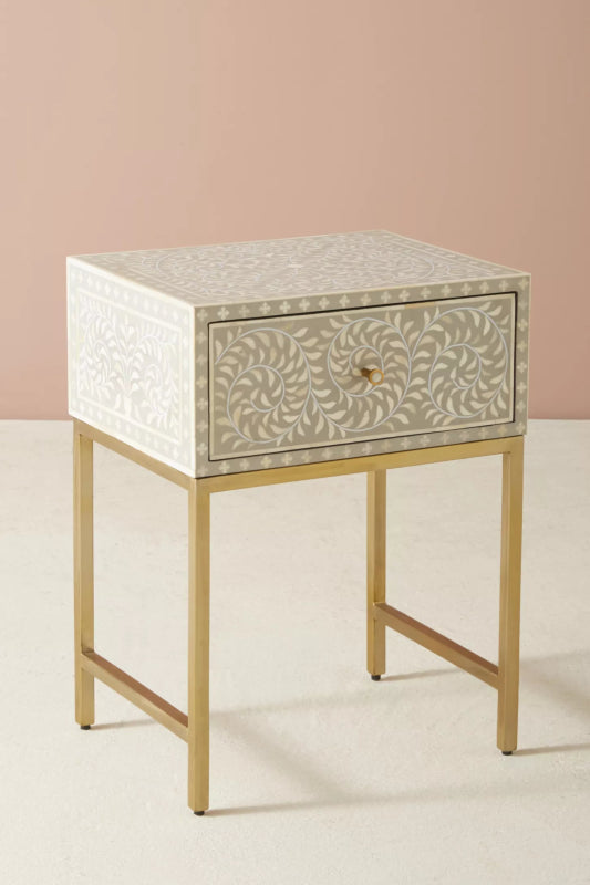 Handmade grey bone inlay nightstand with single drawer storage, all-over floral vine inlay, white dot border on top surface, and brushed gold metal legs on plain background.