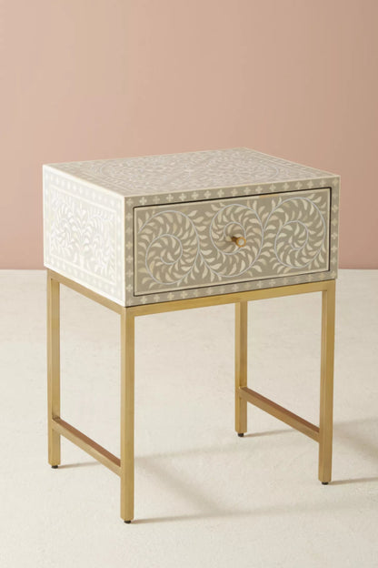 Handmade grey bone inlay nightstand with single drawer storage, all-over floral vine inlay, white dot border on top surface, and brushed gold metal legs on plain background.