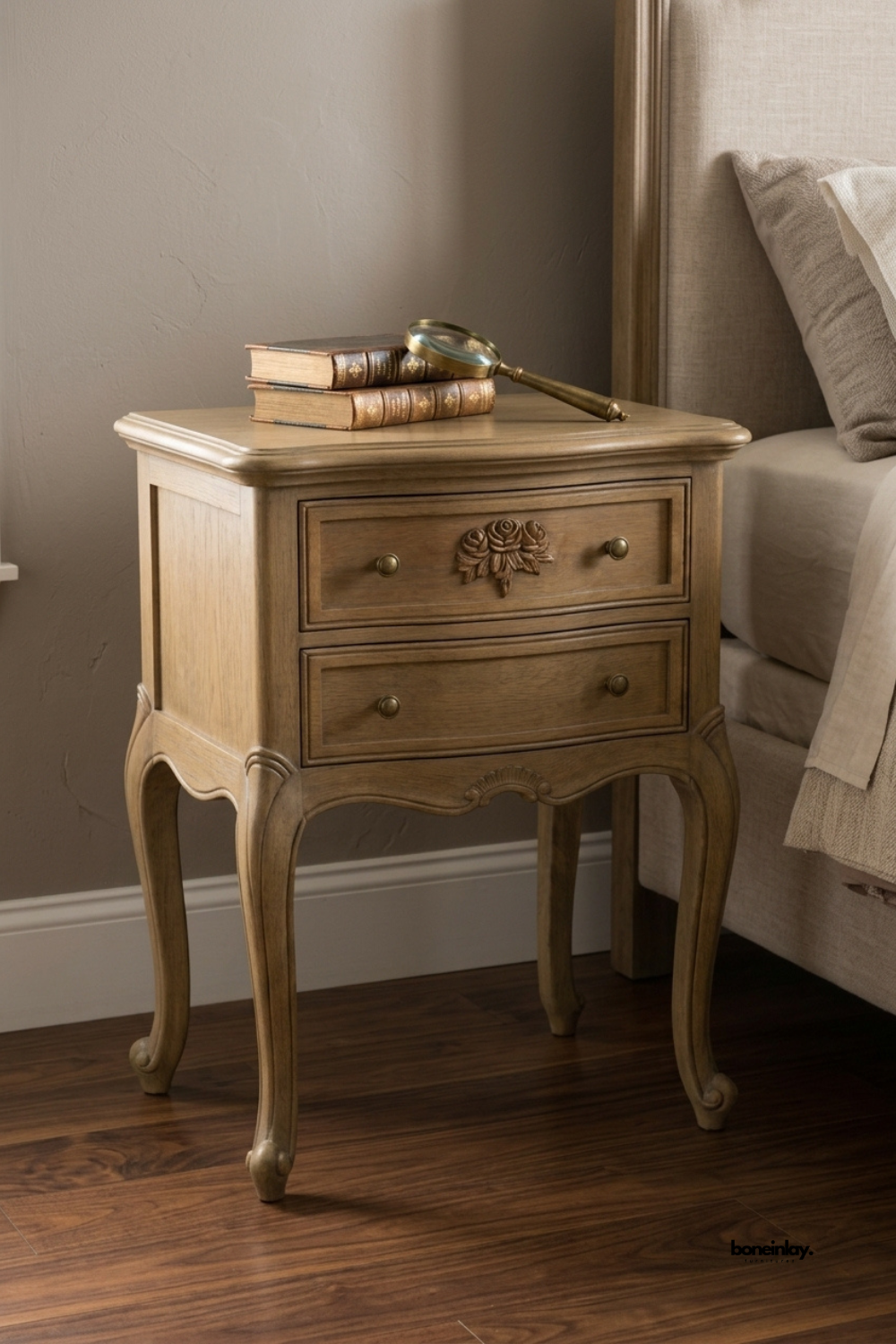 Antique oak nightstand featuring a hand-carved floral rosette drawer front and elegant curved cabriole legs. Two spacious drawers with brass knob pulls offer classic bedside storage. Styled with a stack of leather-bound books and a vintage brass magnifying glass beside a linen bed. A timeless statement nightstand for traditional and vintage-inspired bedrooms.