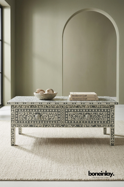 Grey and ivory bone inlay coffee table featuring an intricate floral vine pattern across two front drawers. Slim tapered legs with matching inlay detail complete the elegant silhouette. Styled with a stone bowl of shells and stacked books beneath an arched wall niche. A refined storage coffee table for bohemian and transitional living rooms.
