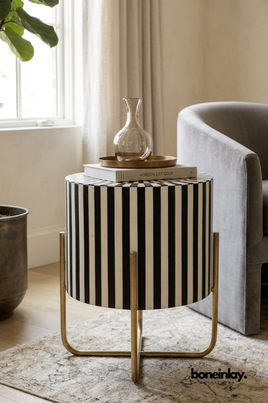 Round bone inlay side table with a bold black and white striped pattern.
Cylindrical drum-shaped top sits on a slim brass X-frame base.
Vertical stripes wrap seamlessly around the entire tabletop surface.
Styled beside a grey armchair with a glass vase and stacked books.