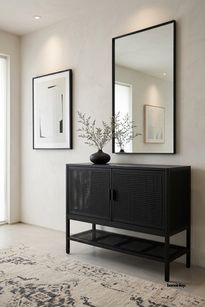 Contemporary matte black 2-door accent cabinet featuring textural woven cane panels, minimalist metal pull handles, and a slatted lower open display shelf. Styled beneath a large black framed mirror in a bright modern entryway, this chic credenza delivers airy bohemian texture and versatile storage to contemporary interiors.