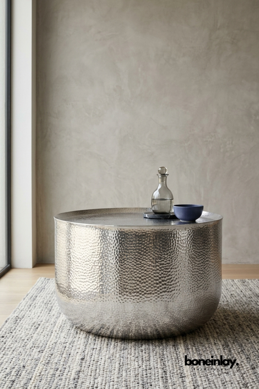 Round hammered silver coffee table featuring a polished nickel finish and sculptural drum silhouette. Dense hammered texture wraps the entire surface for artisanal handcrafted shine. Styled with a glass decanter and a matte navy bowl on a chunky boucle rug. A sleek statement coffee table for modern and contemporary living room interiors.