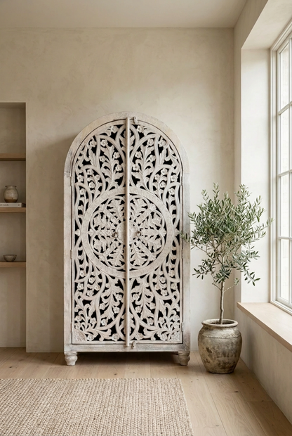 An Indian hand-carved wooden armoire, featuring a front view of doors with intricate floral scrollwork, positioned beside a potted tree and a window.