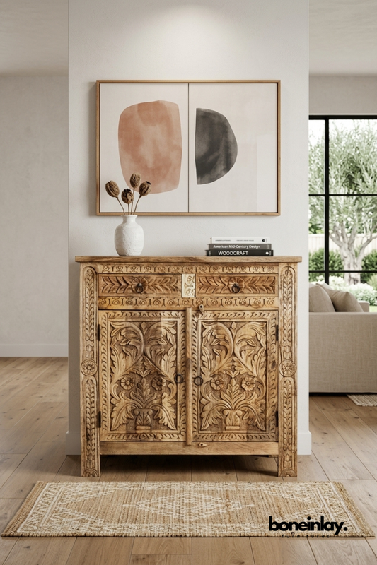 Natural mango wood accent cabinet featuring intricate floral and botanical hand-carved detailing. Includes a top drawer with ring pulls and double carved doors with traditional motifs. Styled with a ceramic vase, stacked books, and framed wall art in a warm neutral setting. A statement storage piece bringing artisanal warmth to entryways and living rooms.