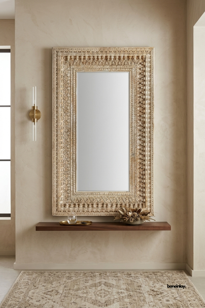 Hand-carved whitewashed wooden wall mirror featuring a multi-tiered carved border with traditional bead and floral relief work. Mounted above a minimalist floating walnut ledge in a warm neutral entryway, this vintage artisan accent mirror brings rustic elegance and timeless craftsmanship to luxury home interiors.