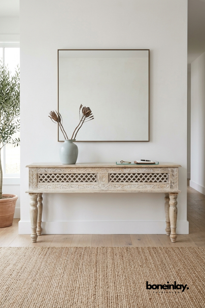 Whitewashed carved wood console table featuring an intricate lattice trellis pattern across two drawer fronts. Floral leaf carvings and turned wooden legs add traditional architectural detail. Styled with a sage ceramic vase, dried botanicals, and a glass tray beneath a square wall mirror. A charming console table for entryways and hallway styling.