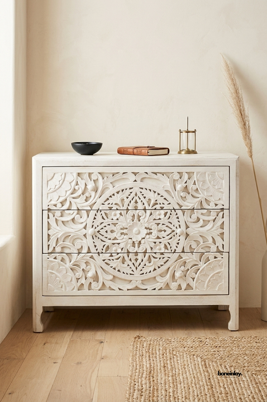 Ivory whitewashed chest of drawers featuring a bold floral medallion pattern carved across three drawer fronts. Scalloped apron and curved bracket feet complete the traditional silhouette. Styled with a matte black bowl, leather book, and brass lantern on a jute rug. A charming statement dresser for bedrooms and bohemian interiors.