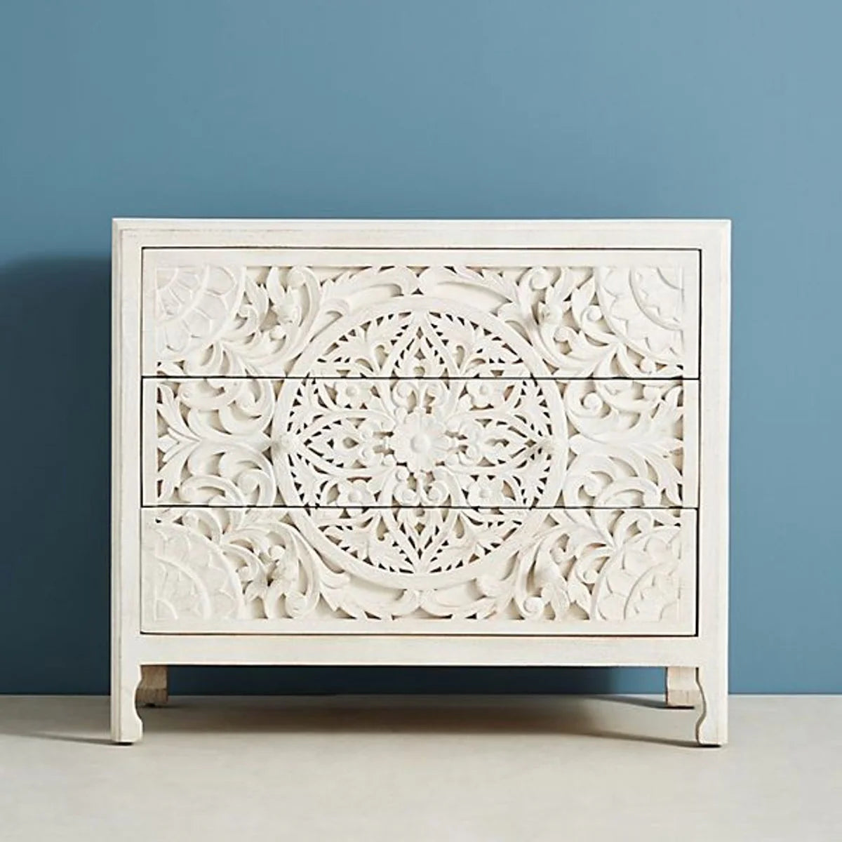 Front view of a hand-carved whitewashed three-drawer wood dresser with an intricate central mandala carving and matching wood knobs.