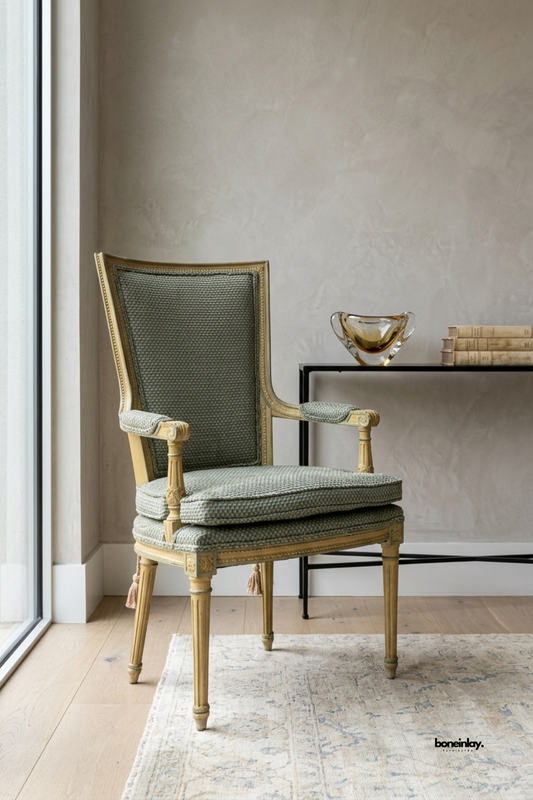Vintage-style armchair featuring a carved and gilded wood frame with tasseled fringe detailing and turned fluted legs. Soft sage green textured upholstery covers the tall back, seat cushion, and padded armrests. Styled beside a black metal side table with an amber glass vase and stacked books. An elegant statement armchair for formal living rooms and reading nooks.