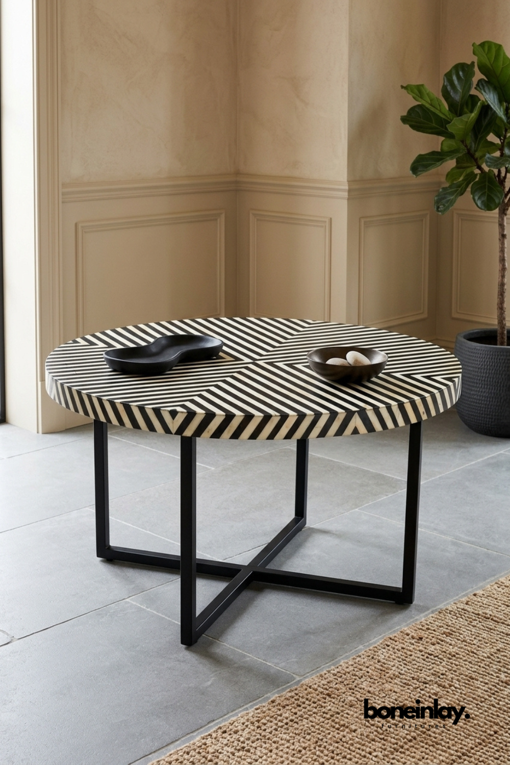 Round bone inlay coffee table featuring a bold pinwheel chevron pattern in contrasting black and white stripes. Sleek black crossed metal legs add a modern architectural base to the graphic tabletop. Styled with matte black bowls holding decorative stones on a neutral stone floor. A striking statement coffee table for contemporary living spaces.