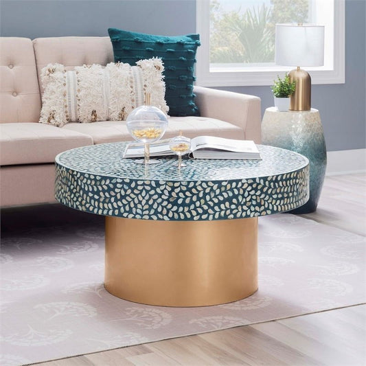 A round coffee table with a teal resin top and shimmering white leaf-patterned Mother of Pearl inlay, resting on a tall cylindrical gold brass base.