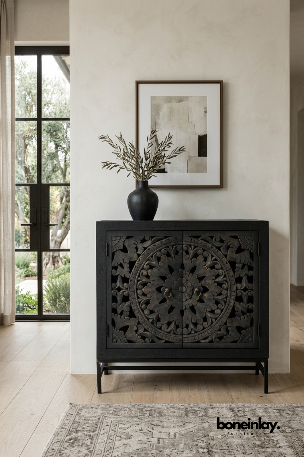Matte black carved wood accent cabinet featuring a bold floral mandala pattern on double doors. Set on a slim black metal base for a modern silhouette with traditional carving detail. Styled with a black ceramic vase and olive branches beneath framed abstract art. A dramatic accent cabinet perfect for entryways and statement wall vignettes.