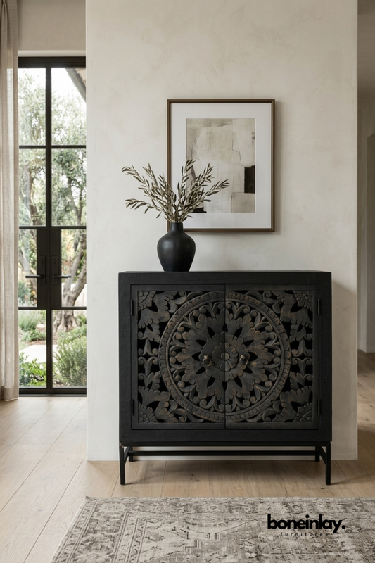 Matte black carved wood accent cabinet featuring a bold floral mandala pattern on double doors. Set on a slim black metal base for a modern silhouette with traditional carving detail. Styled with a black ceramic vase and olive branches beneath framed abstract art. A dramatic accent cabinet perfect for entryways and statement wall vignettes.