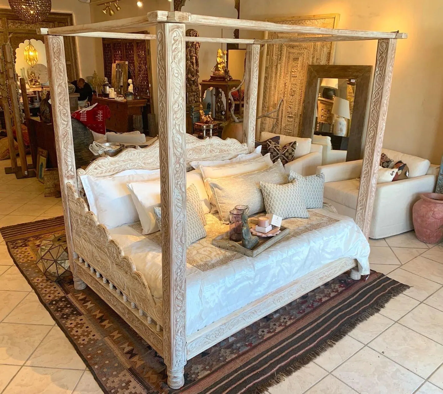 A luxury hand-carved solid wood four-poster canopy daybed with a whitewash finish, intricate vine carvings on pillars, and a scalloped frame styled with white and beige linens.