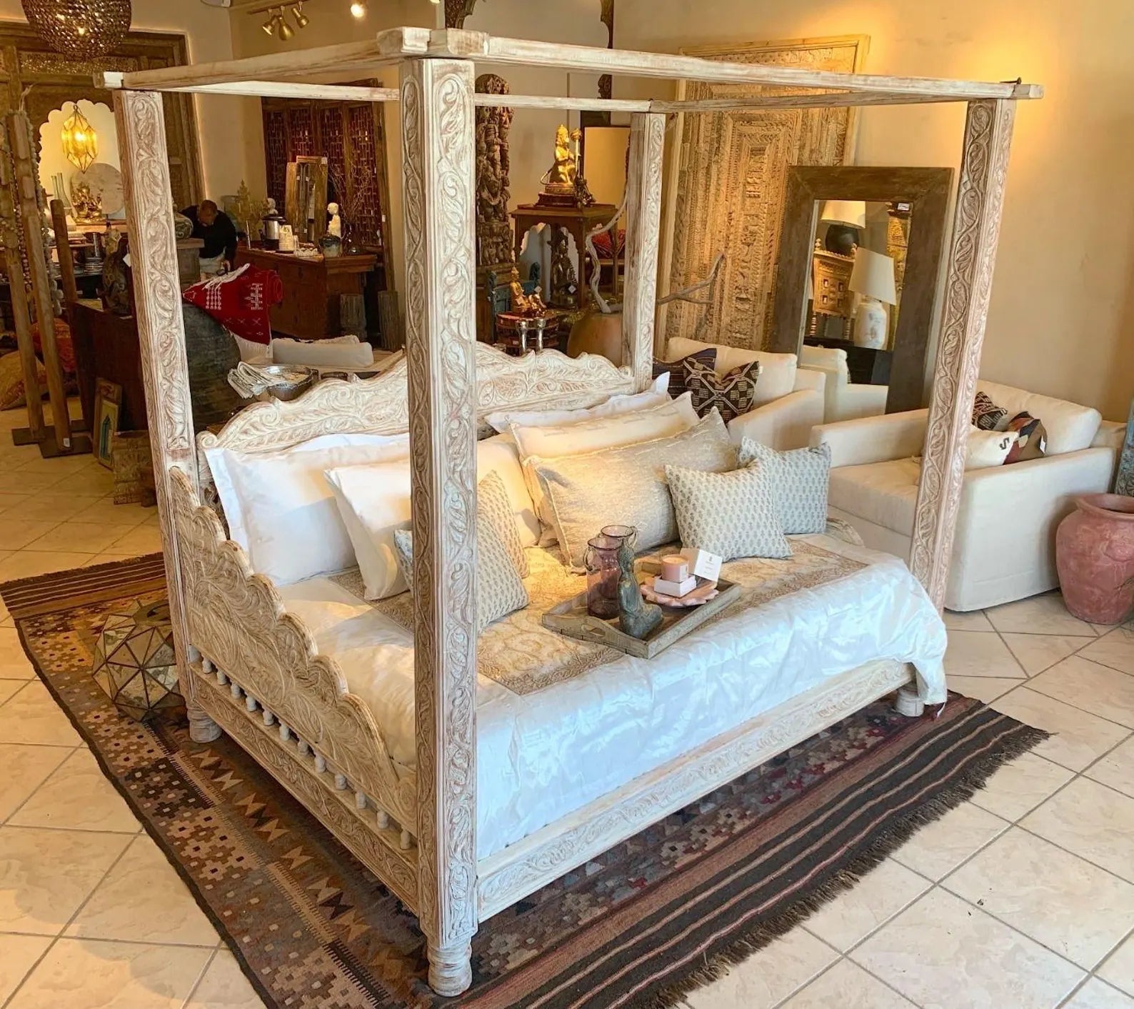 A luxury hand-carved solid wood four-poster canopy daybed with a whitewash finish, intricate vine carvings on pillars, and a scalloped frame styled with white and beige linens.