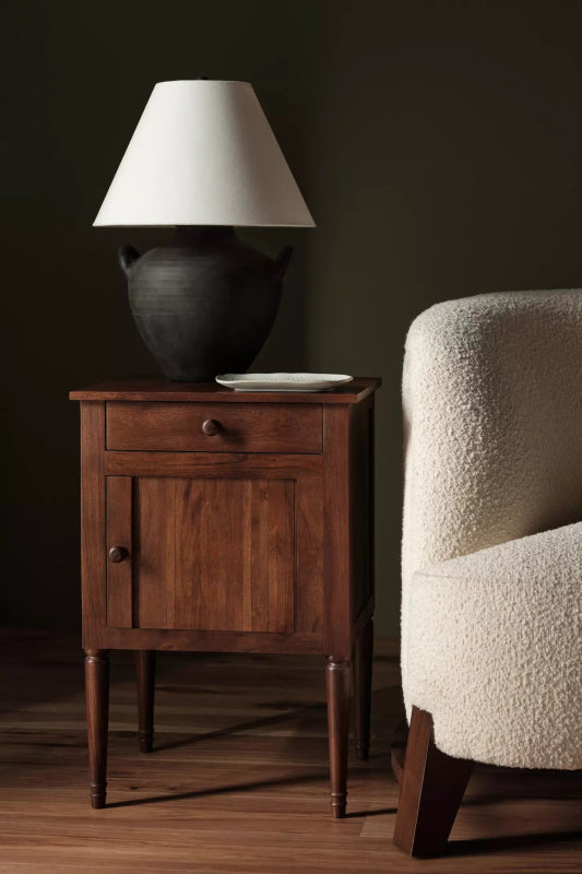 Natural finish wooden bedside table with one drawer and one door, styled with a lamp beside a cream armchair