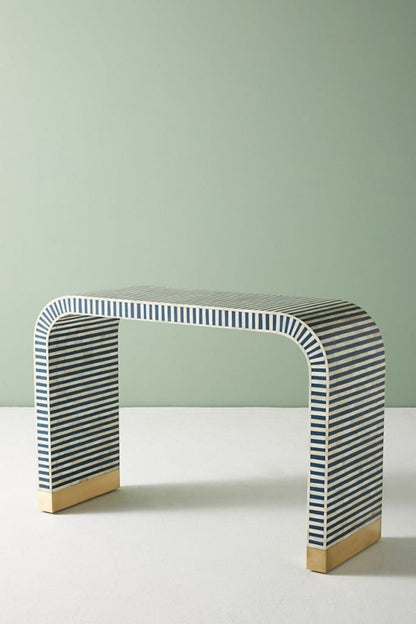 Three-quarter side angle of the blue bone inlay entryway table, showing the continuous waterfall curve, striped inlay surface, slim console profile, and brass-capped feet. the handmade table as a narrow hallway.