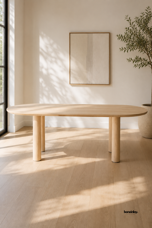 Solid light oak oval dining table featuring thick cylindrical pillar legs and a smooth rounded wooden tabletop. Styled in a sunlit minimalist dining room with neutral walls and a potted olive tree, this artisan wooden table brings Scandinavian modern design and organic warmth to luxury home interiors.