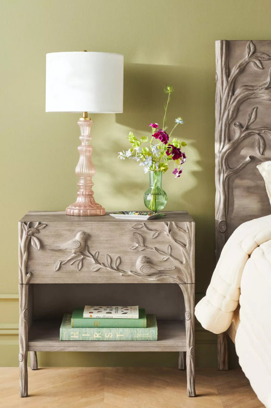 Natural bird carved nightstand in solid wood, styled in bedroom beside a bed with a lamp, flowers and shelf books.