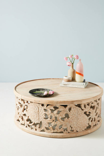 Natural round center table in carved wood, front view with pink flowers, decorative bottles and a black dish.