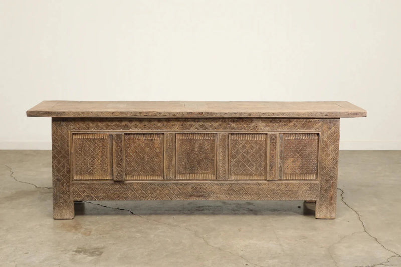 Natural wood sideboard with carved door fronts and a long overhanging top, straight-on view against a pale wall.