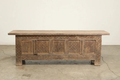 Natural wood sideboard with carved door fronts and a long overhanging top, straight-on view against a pale wall.