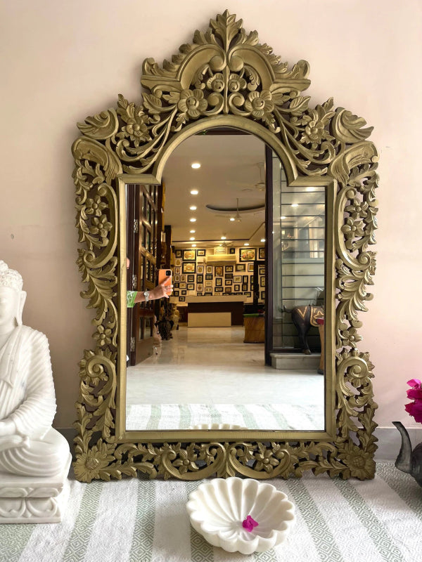 Ornate arched wall mirror in gold-toned carved wood, front view beside a white figurine and decorative bowl.