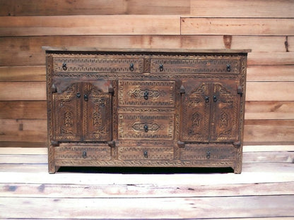 Ornate four door cupboard in rustic carved wood, front view showing closed drawers against a horizontal plank wall.