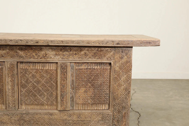 Paneled wood cabinet in a weathered brown finish with carved door fronts, closer front view against a pale wall.