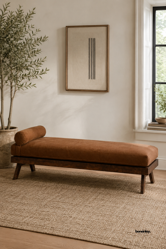 Handmade wooden daybed featuring a rich dark wood frame, angled support legs, and a plush brown upholstered cushion complete with a cylindrical bolster pillow. Styled in a warm, minimalist room interior with an olive tree and natural jute rug, this artisan lounge piece brings mid-century modern elegance and cozy comfort to contemporary US homes.