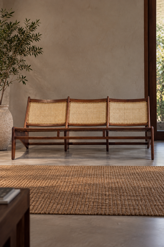 Pierre Jeanneret inspired three-seater wooden bench with three woven cane backrests, a dark wooden frame and angular legs, shown from the front against a neutral wall, with a potted tree beside it and a textured rug in the foreground.