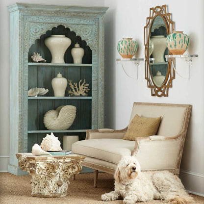 Rustic floral carved bookshelf in solid wood, styled in living room with ceramic jars and shells beside an armchair.
