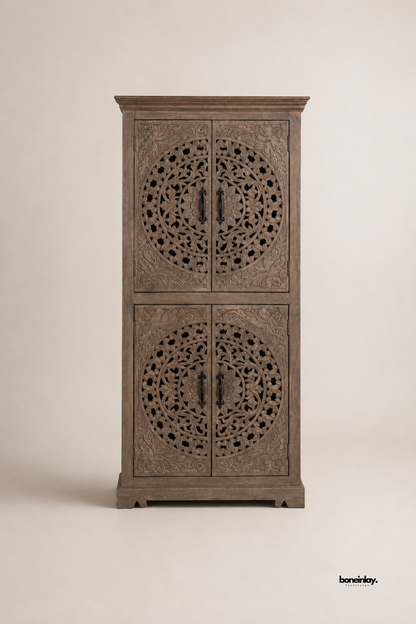 Grey-washed wood bar cabinet featuring intricate circular floral medallion carvings across four cabinet doors. Black metal handles and a crown molding top add refined architectural detail to the tall silhouette. Photographed on a plain studio background to showcase the dense hand-carved craftsmanship. A striking statement bar cabinet for dining rooms and entertaining spaces.