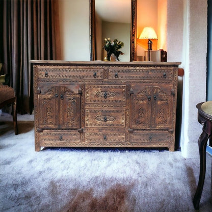 Rustic seven drawer server in carved wood, styled in living room with a tall mirror, glowing table lamp and flowers.