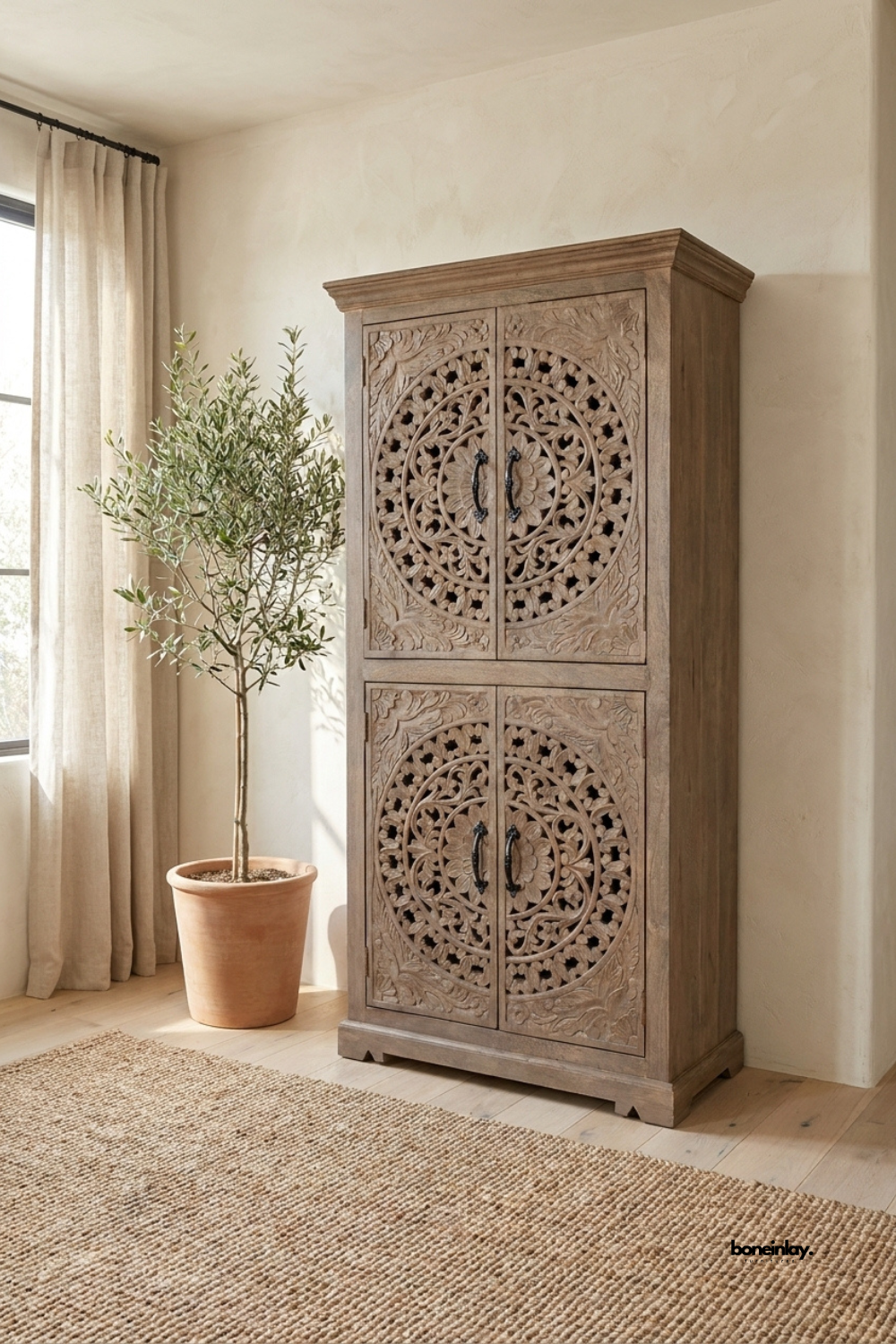 Weathered grey-brown wood Armoire featuring intricate circular floral medallion carvings across four cabinet doors. Black metal handles and a crown molding top complete the tall, architectural silhouette. Styled beside a terracotta planter and olive tree on a jute rug near a curtained window.
