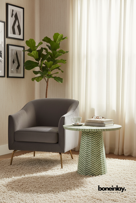 Sage green and ivory chevron bone inlay pedestal side table styled in a modern living room beside a grey velvet armchair on a cream rug.
