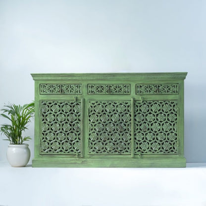 Sage green floral sideboard in solid wood, front view showing three carved doors and three drawers beside a potted plant.