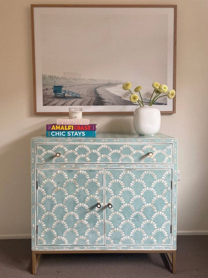 Seafoam blue and white bone inlay scalloped cabinet with one drawer and two doors styled on a wood floor with stacked books, candle, and white vase with yellow flowers