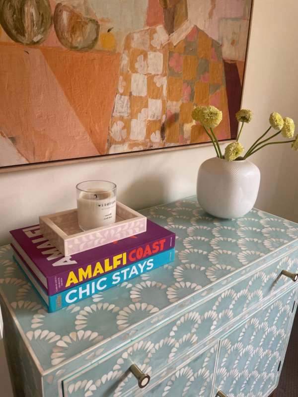 Seafoam blue bone inlay scalloped cabinet with books, candle tray, and white ceramic vase with yellow flowers on top, abstract artwork hanging above on a neutral wall