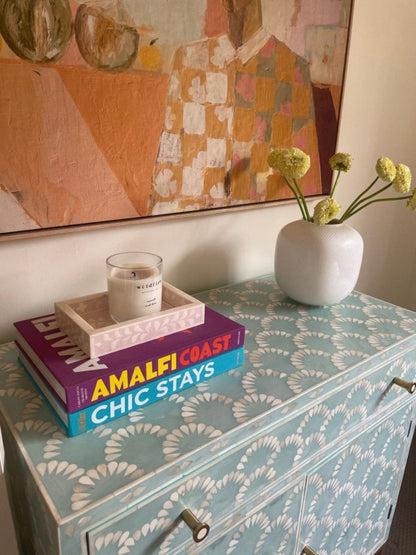 Seafoam blue bone inlay scalloped cabinet with books, candle tray, and white ceramic vase with yellow flowers on top, abstract artwork hanging above on a neutral wall
