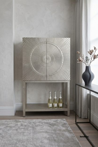 Artisan-crafted tall bar cabinet showcasing double doors with a dimensional carved radial sunburst medallion in an antique silver champagne metal finish over an open plinth base. Styled against soft grey plaster walls on a plush area rug, this luxury statement credenza brings glamorous Art Deco flair and elevated entertaining storage to modern living spaces.
