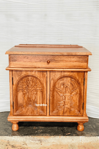 Solid wood storage cabinet in warm natural wood, front view showing a single drawer, carved double doors and turned feet.