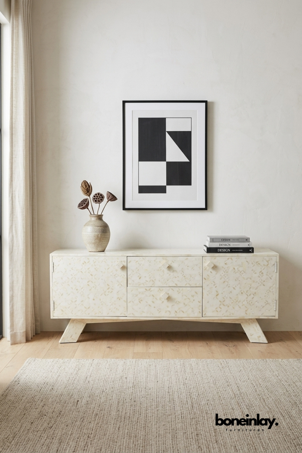 Ivory bone inlay media console featuring a floral mosaic pattern, center drawers, and angled wooden legs. Styled with a stoneware vase, dried botanicals, and stacked design books beneath framed abstract art. Sleek silhouette blends bohemian texture with mid-century modern lines. An elegant storage solution for living rooms and dining spaces.